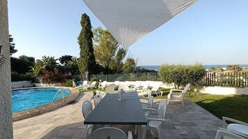 An outdoor pool area with a table and chairs, surrounded by a white fence and greenery.