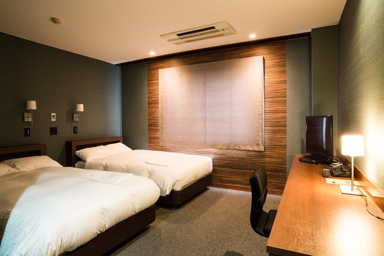 A hotel room with two beds, a desk with a computer and phone, a TV, and a large window with blinds.