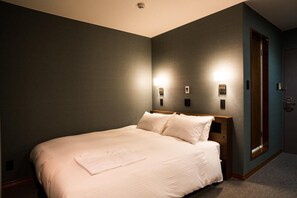 Una cama cuidadosamente tendida en una habitación de hotel, con cabecera de madera, dos lámparas fijadas a la pared y una puerta a la derecha.