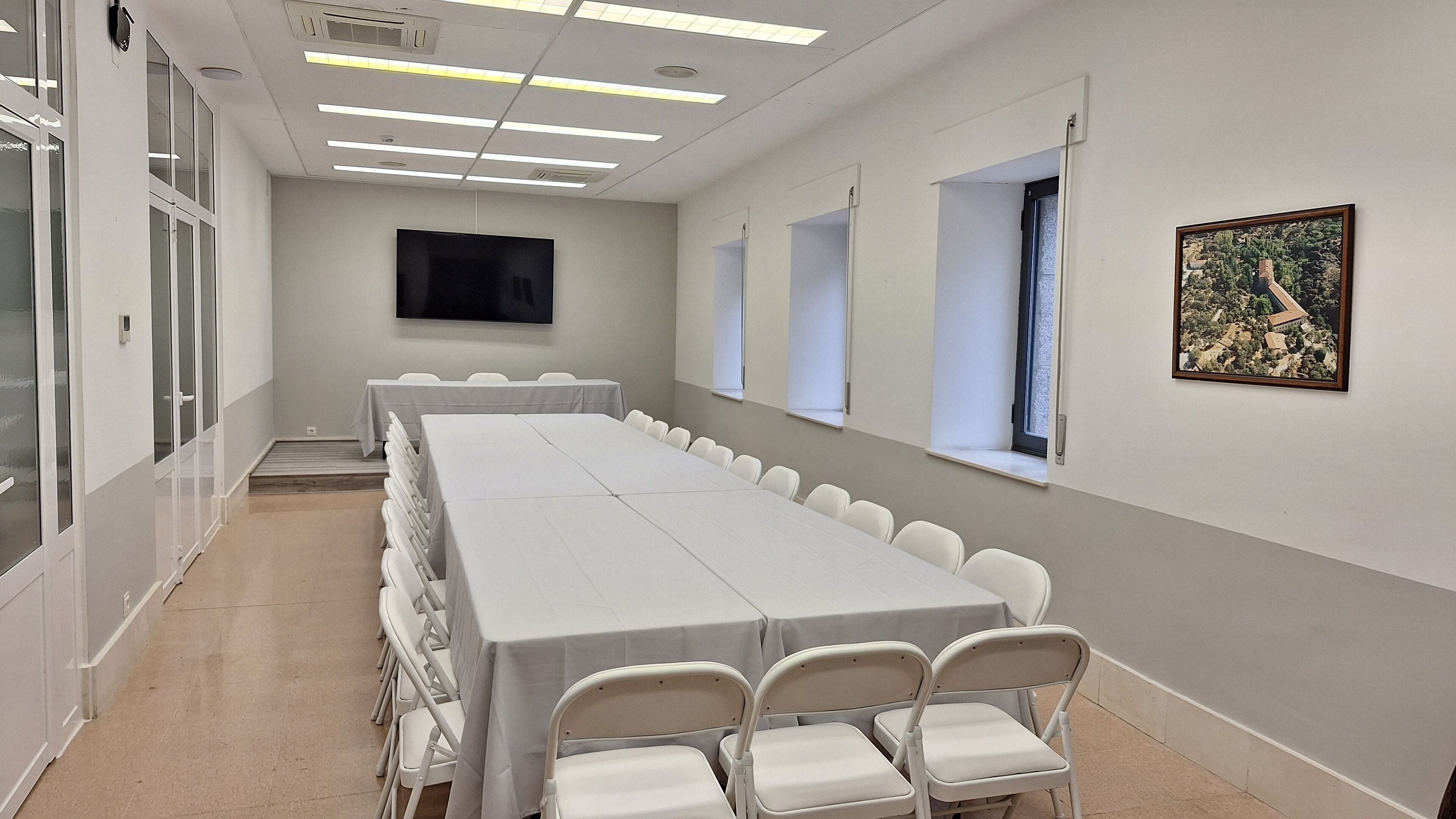 Un salón de conferencias con una larga mesa, sillas blancas, un televisor de pantalla plana, ventanas y un cuadro en la pared.