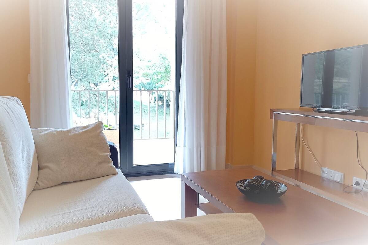 A living room with a sofa, a television on a stand, and a view of a balcony with greenery.