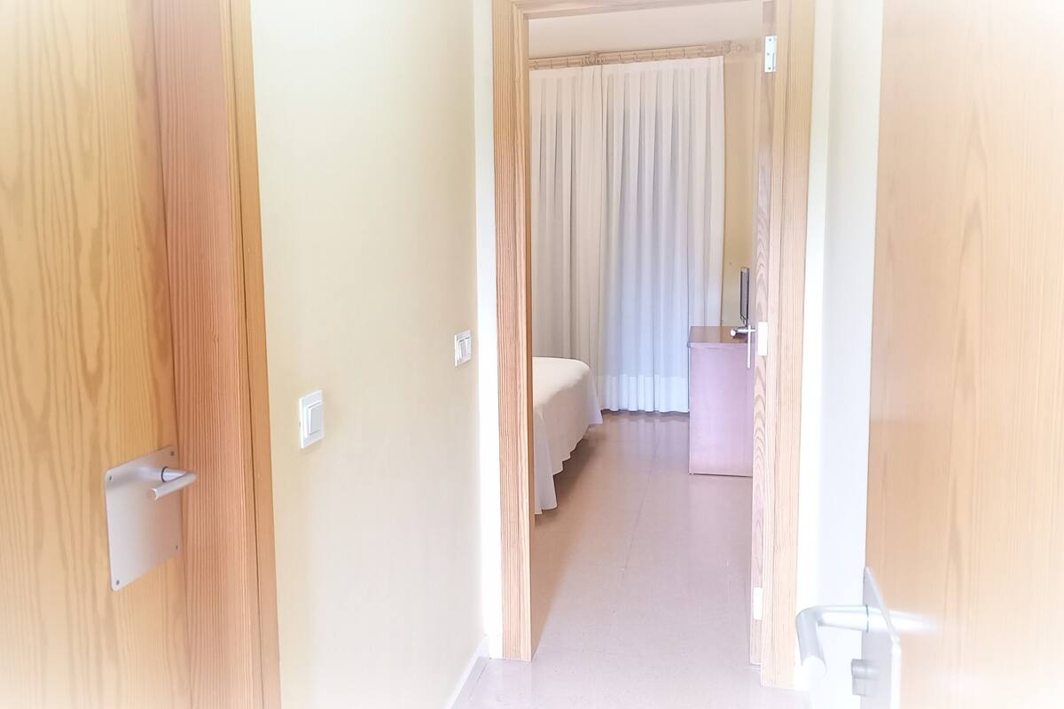 A hotel room with a wooden door, a bed, a small table, and a window with curtains.