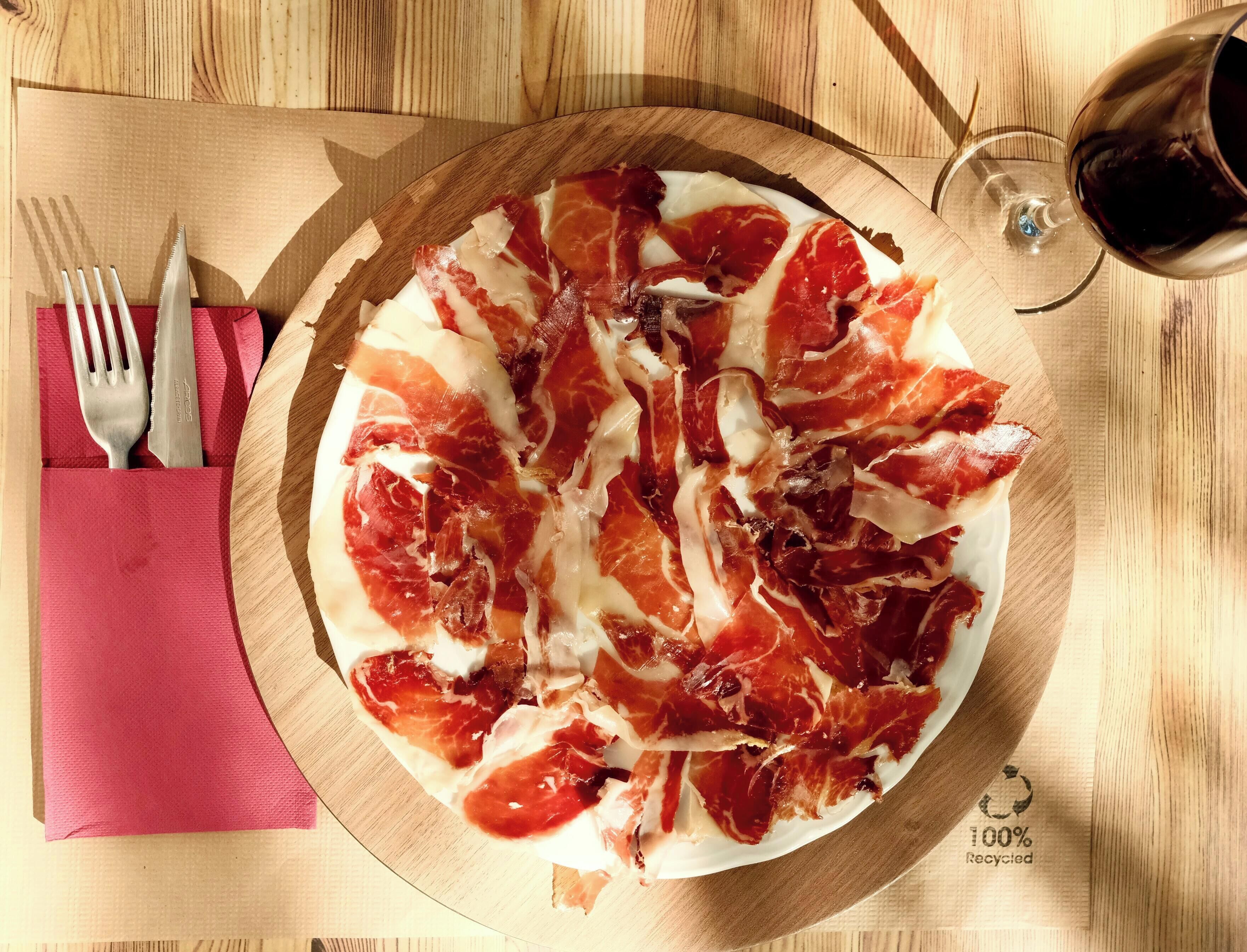 Pizza amb pernil salat sobre un tauler de fusta, un got de vi negre i un gots amb coberts sobre una tovallola vermella damunt una taula de fusta.