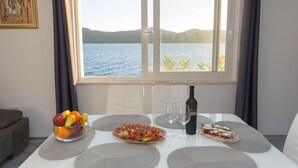 Una mesa de comedor con frutero, platos de comida, copas de vino y una botella, frente a una ventana con vista a un lago y colinas.