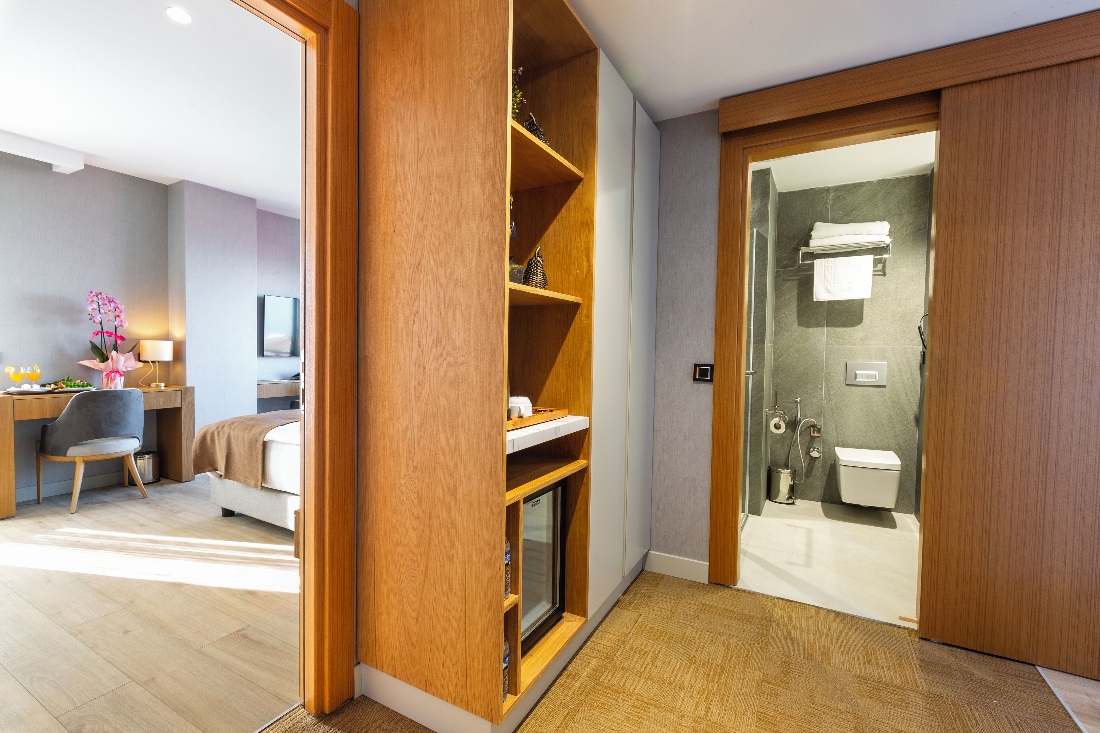A modern hotel room with a wooden wardrobe, a desk with a chair, a bed, and a bathroom with a toilet and shower.