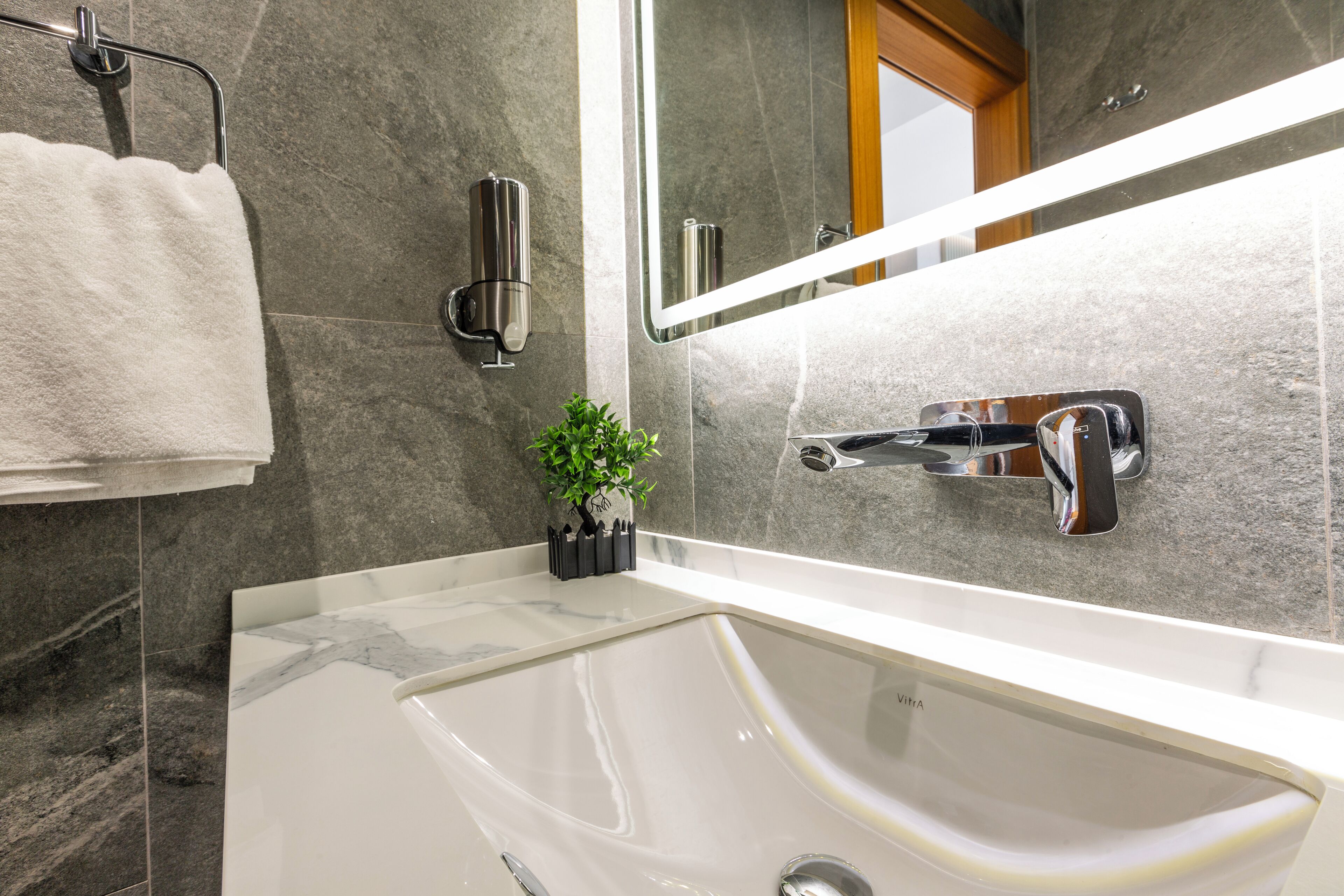A modern bathroom with a large sink, a towel rack, a wall-mounted mirror, and a small potted plant.