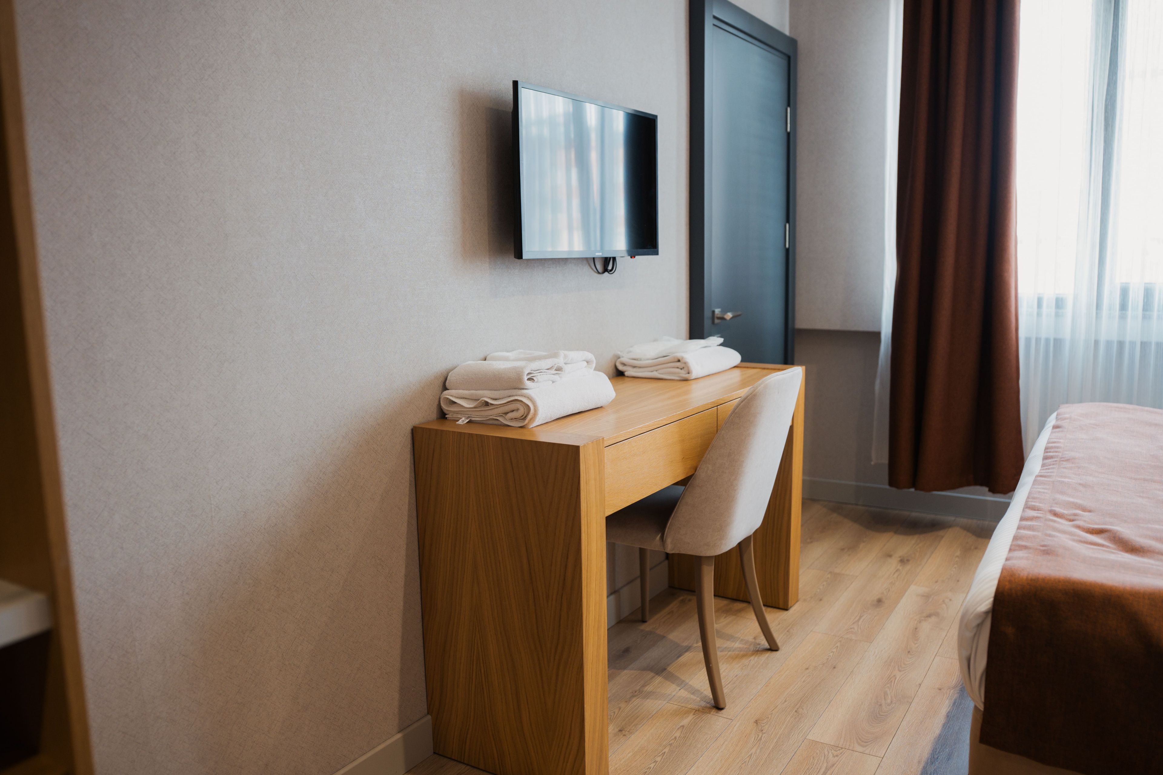 A hotel room with a wooden desk, a chair, a bed, a television mounted on the wall, and a door.