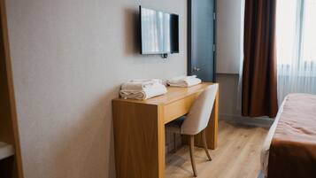 A hotel room with a wooden desk, a chair, a bed, a television mounted on the wall, and a door.
