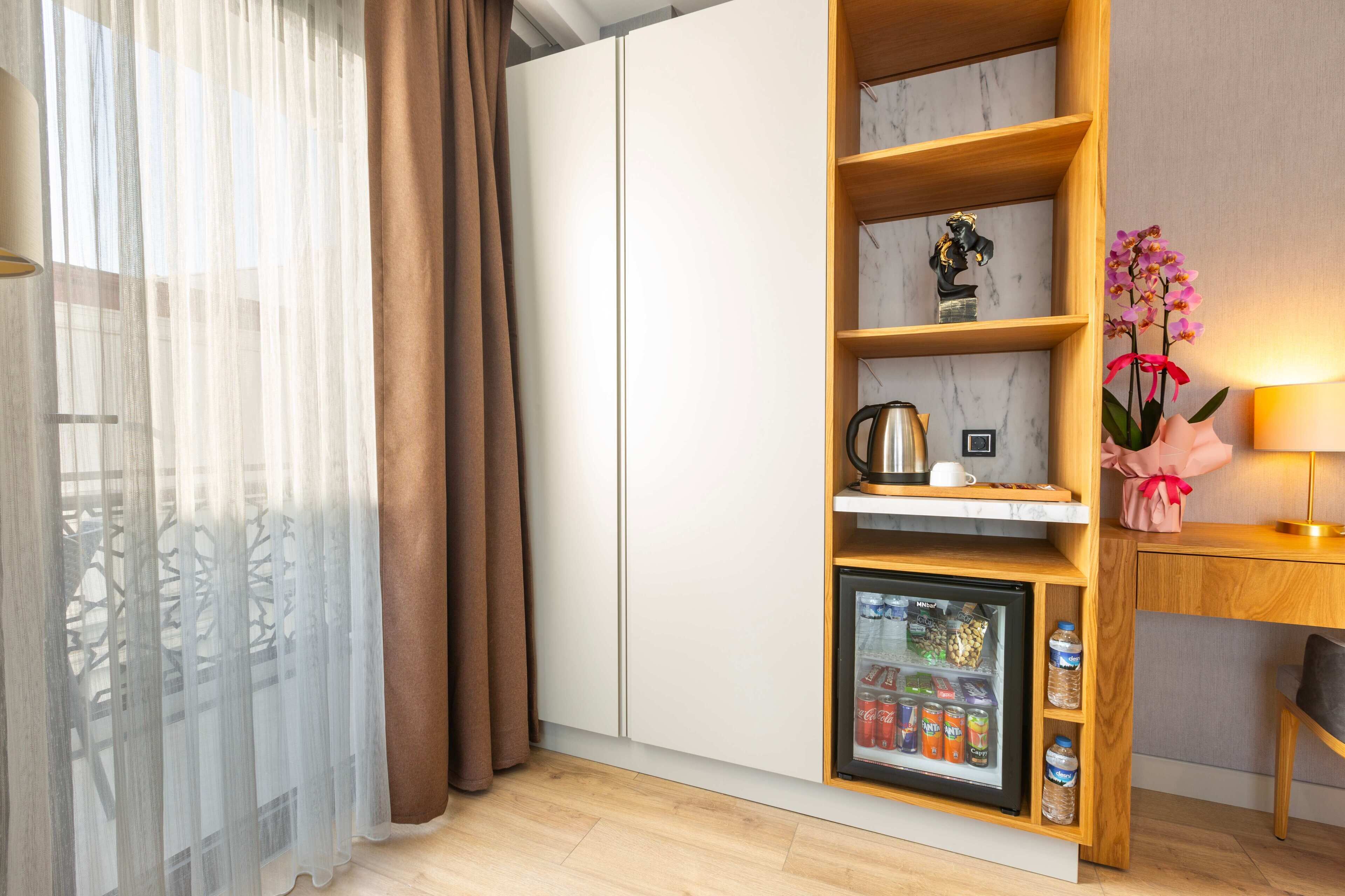 A hotel room with a minibar, a wooden shelf with decorative items, a small refrigerator, and a window with sheer curtains.