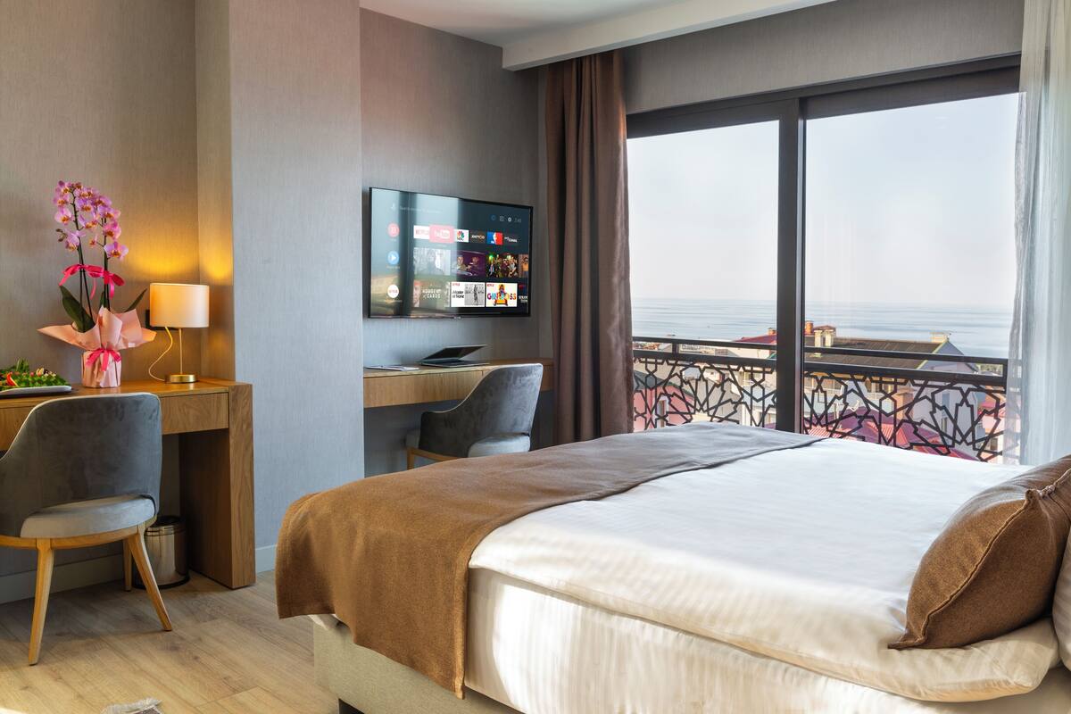 A hotel room with a bed, desk, chair, TV, and a balcony with a view of the sea.