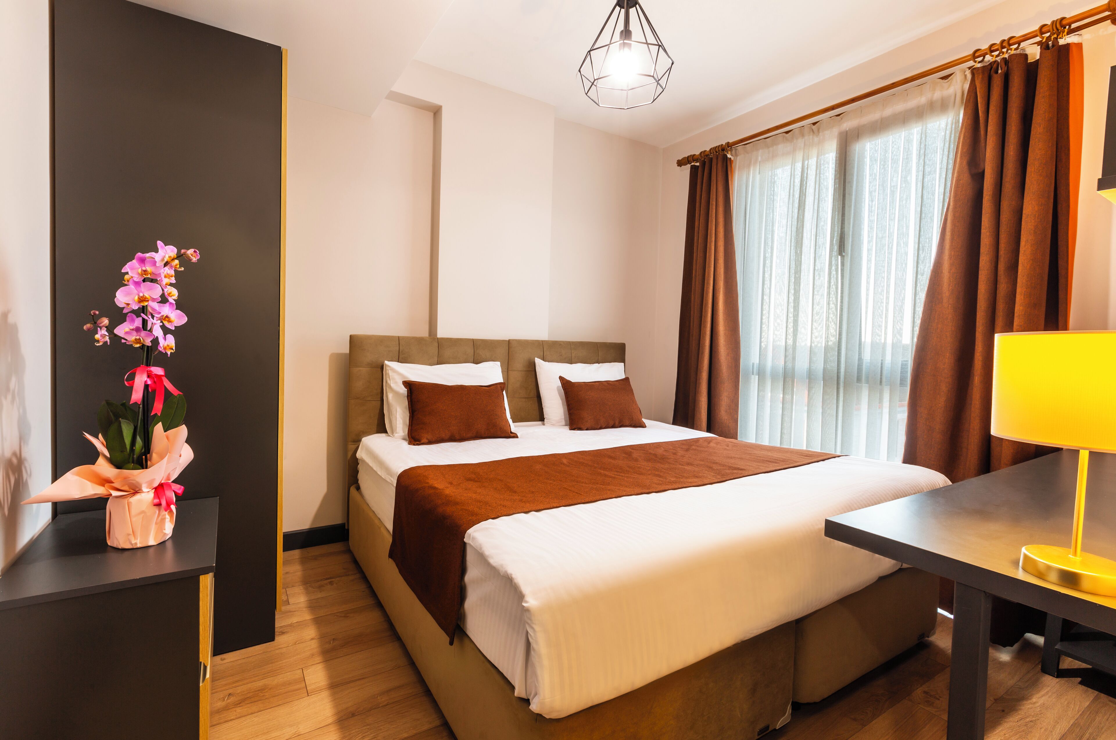 A hotel room with a bed, a bedside table, a lamp, a vase with flowers, and a window with curtains.