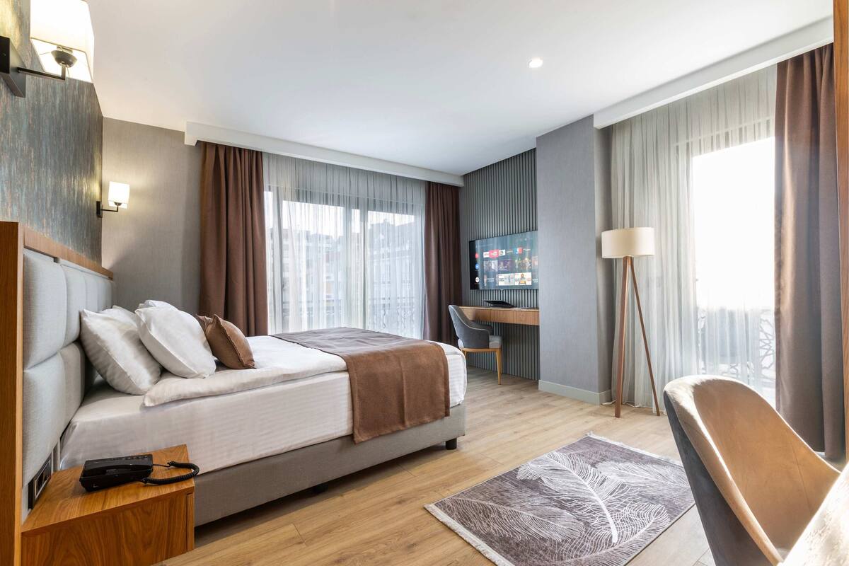 A modern hotel room with a bed, desk, chair, and a large window with curtains.
