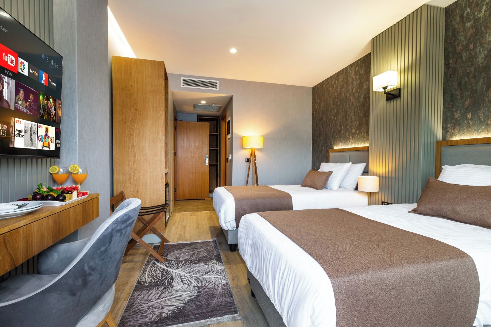 Habitación de hotel con dos camas, un escritorio con silla, un televisor montado en la pared y una pequeña mesa de comedor con frutas y bebidas.