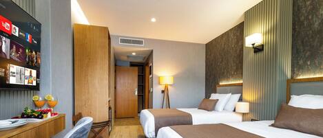 Habitación de hotel con dos camas, un escritorio con silla, un televisor montado en la pared y una pequeña mesa de comedor con frutas y bebidas.