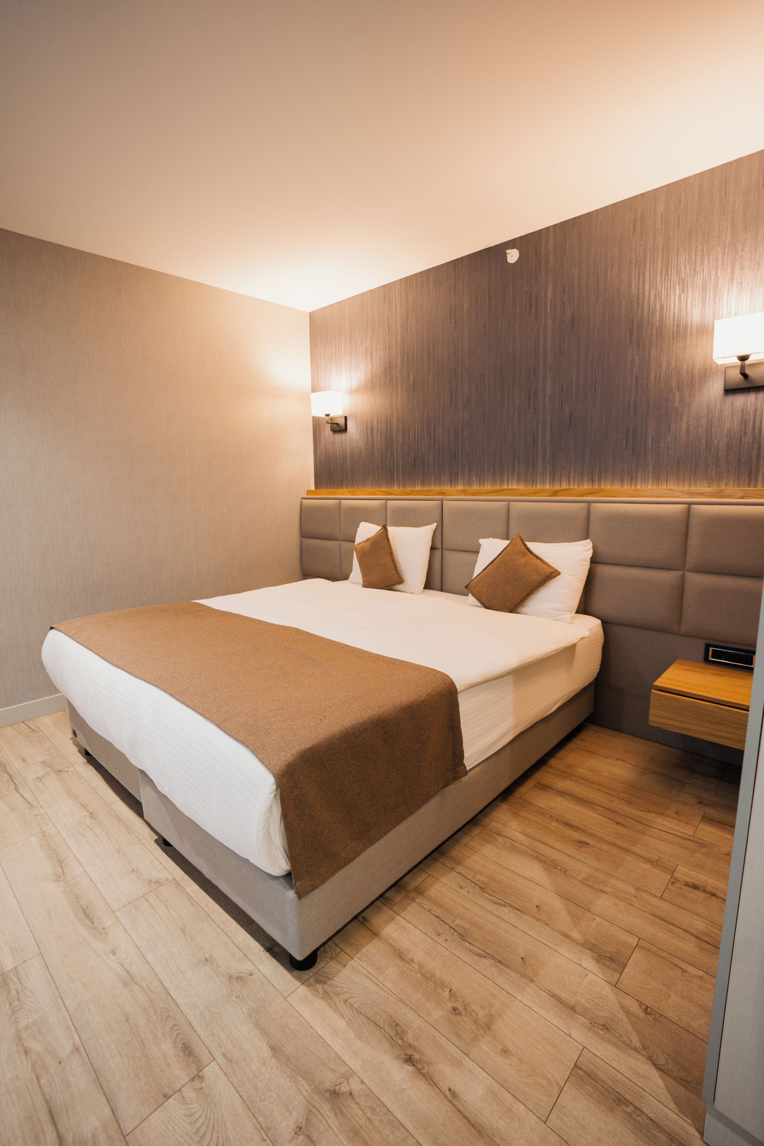 A modern hotel room with a large bed, a wooden headboard, a nightstand, and a wall-mounted lamp.