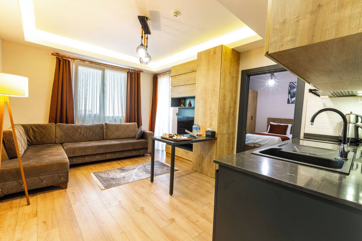 A compact living space with a sofa, a small dining table, a kitchen area with a sink, and a bed in the background.