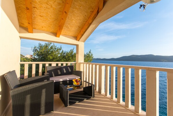 A balcony with wicker furniture and a fruit bowl, overlooking a body of water and distant mountains.