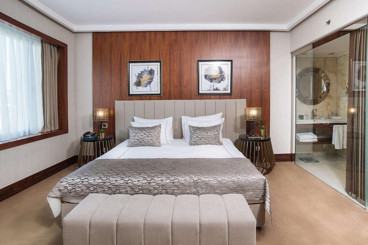 A hotel room with a large bed, two bedside tables with lamps, a wooden headboard, and two framed artworks on the wall.