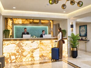 A hotel reception desk with marble countertops, two receptionists, a guest with a suitcase, and modern decor including a chandelier and artwork. - Sura Luxury Million & Spa (Istanbul)