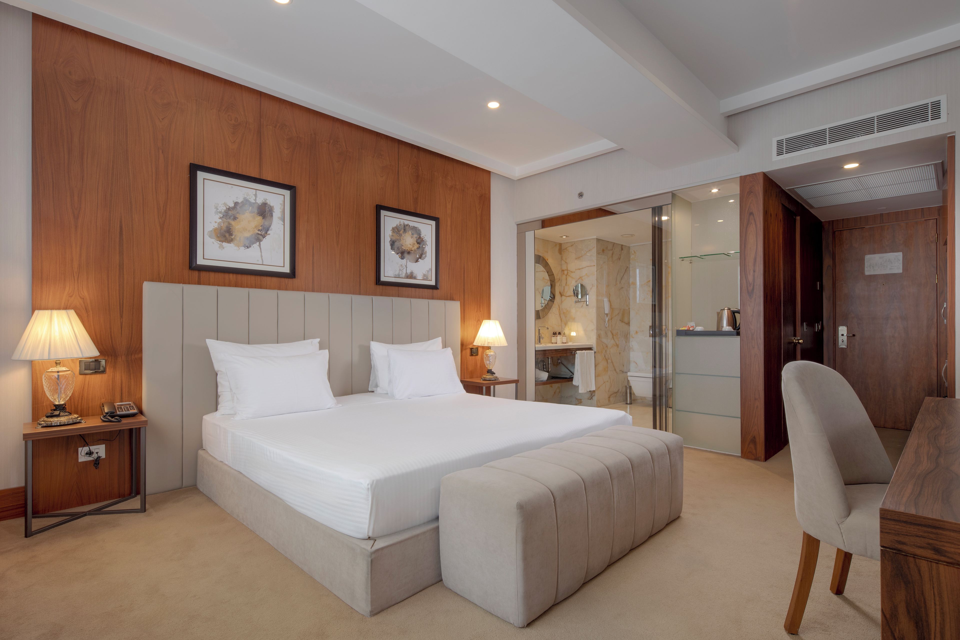 Una habitación de hotel con una cama grande, lámparas de noche, un cabecero de madera, un escritorio con una silla y un baño visible a través de una puerta de cristal.