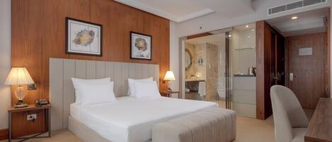Una habitación de hotel con una cama grande, lámparas de noche, un cabecero de madera, un escritorio con una silla y un baño visible a través de una puerta de cristal.