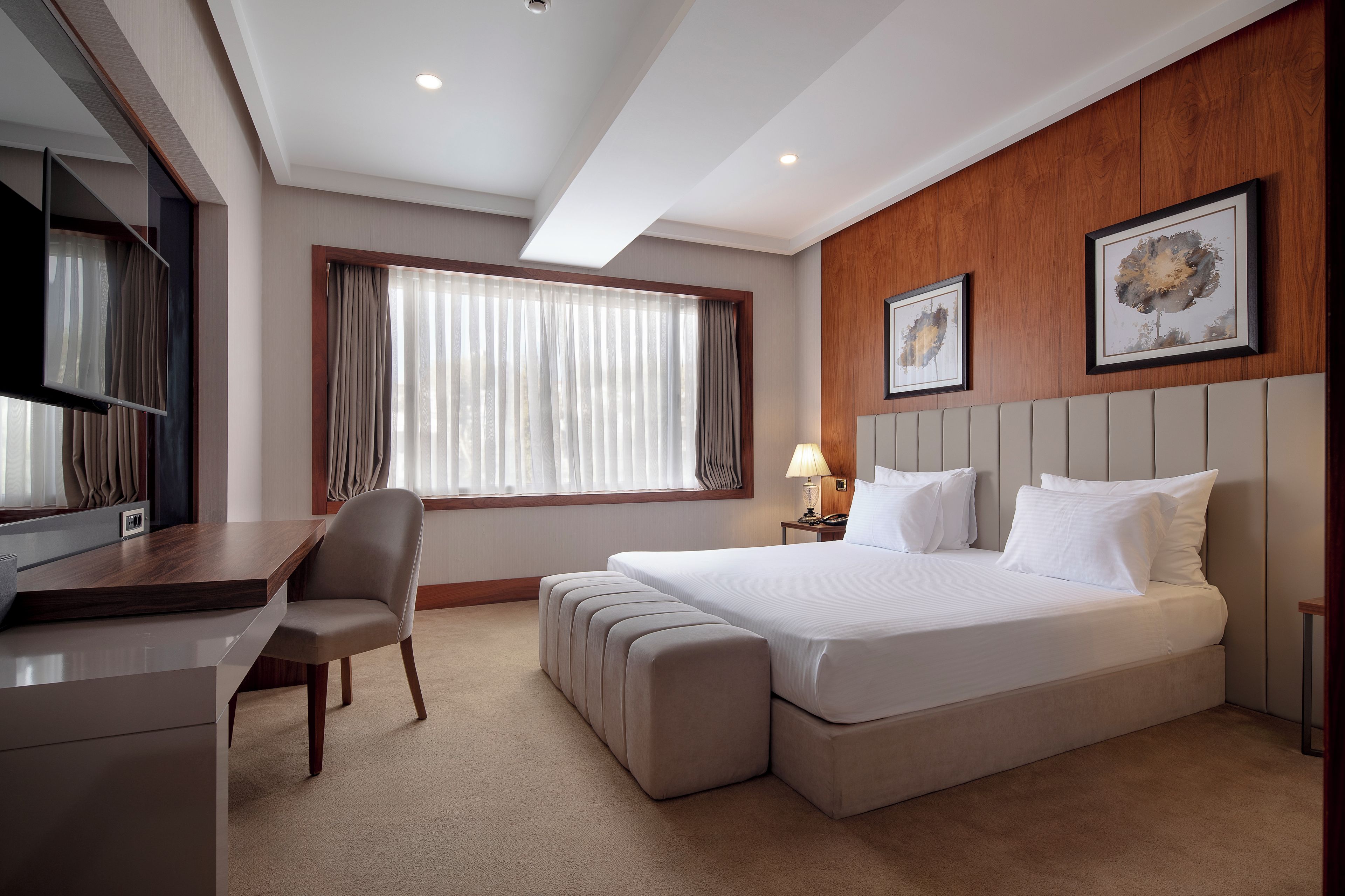 Habitación de hotel con una cama, un escritorio, una silla, una lámpara y dos cuadros en la pared.