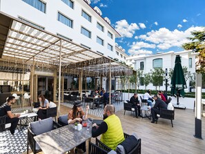 An outdoor cafe area with tables and chairs, a covered seating area with a wooden ceiling, and a building with multiple windows in the background. - Sura Luxury Milion & Spa (Istanbul)