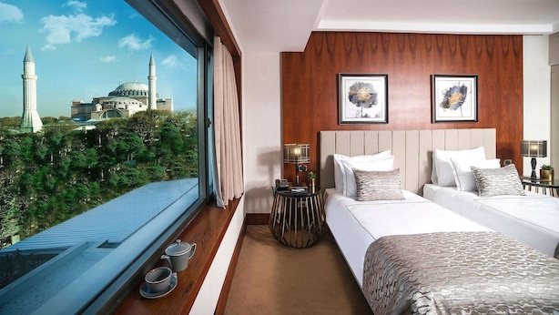 A hotel room with a large window offering a view of a mosque and minaret, a bed with a headboard, bedside tables with lamps, and a small round table with a plant. - Sura Luxury Million & Spa (Istanbul)