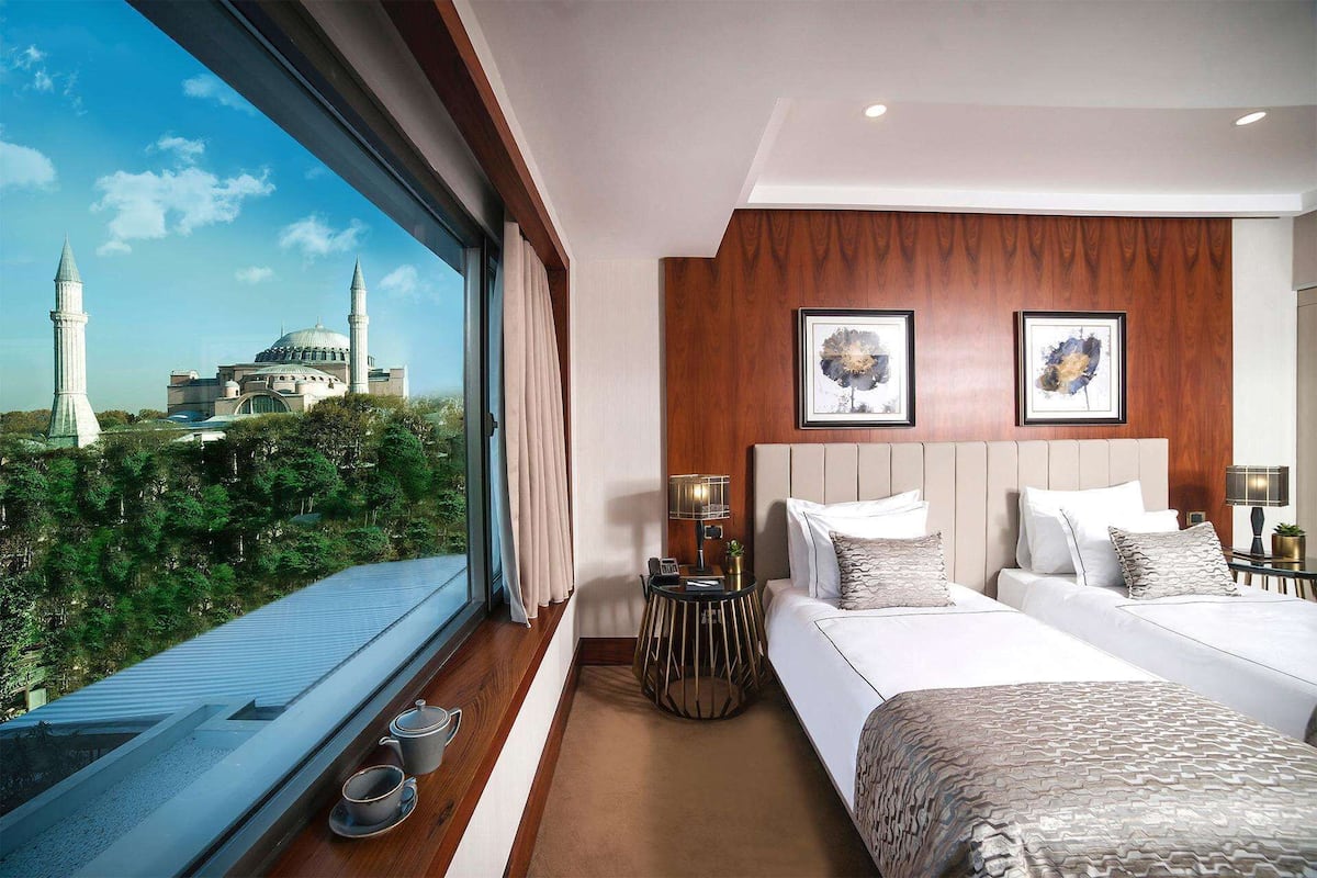 A hotel room with a large window offering a view of a mosque and minaret, a bed with a headboard, bedside tables with lamps, and a small round table with a plant.
