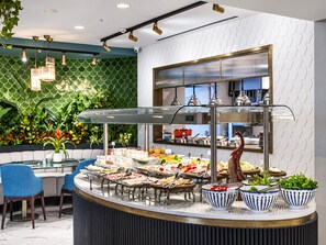 Montaje de bufé con diversas opciones de comida, un área de asientos con sillas y una pared decorativa con diseño geométrico.