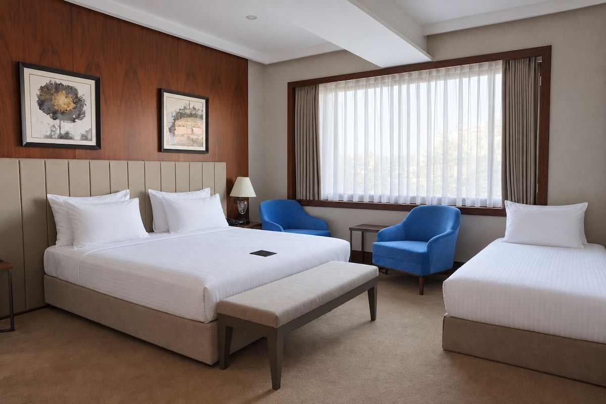 A hotel room with a large bed, two single beds, a blue armchair, a small table, and a window with curtains.