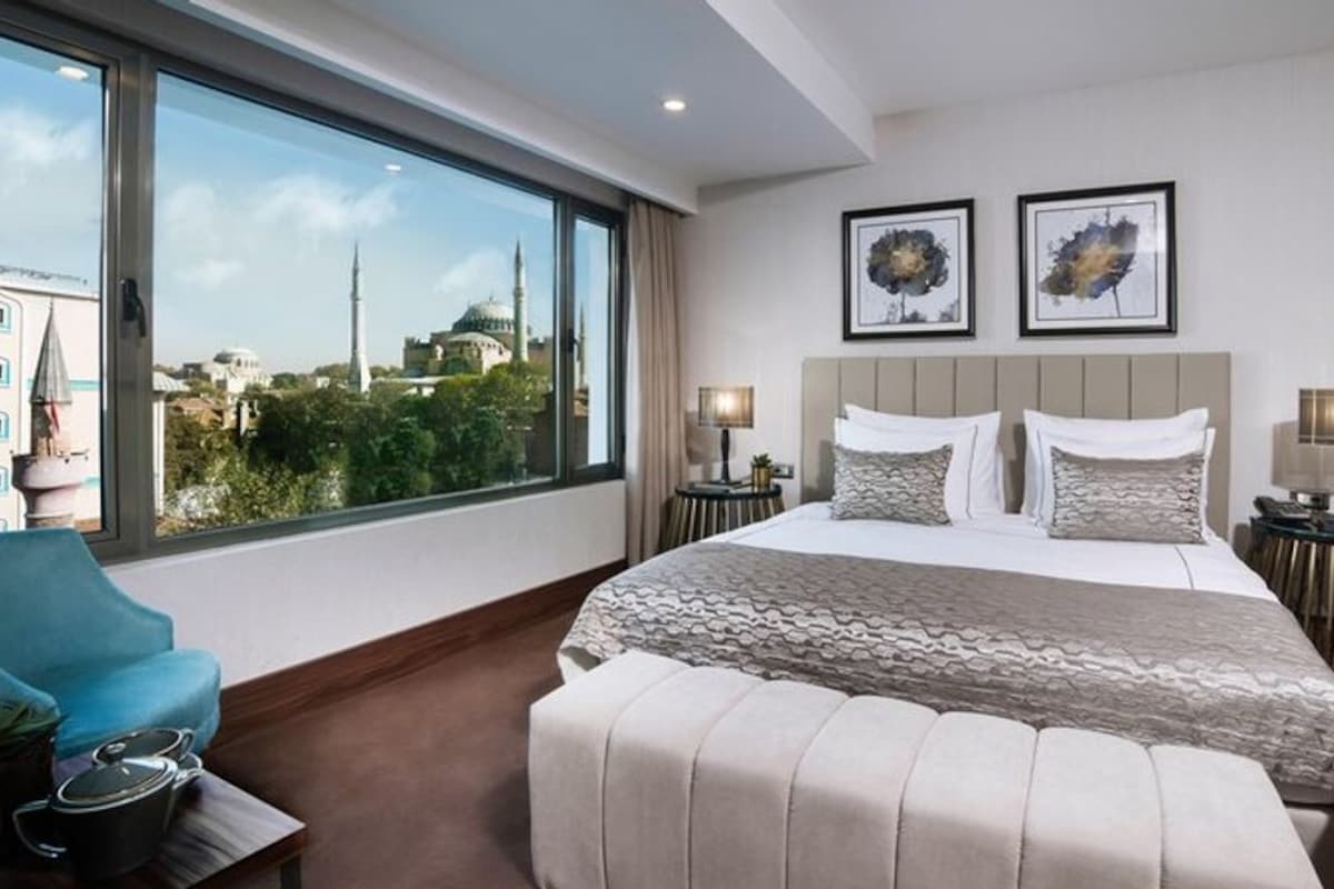 A hotel room with a large bed, two bedside tables, a blue armchair, and a view of a mosque and buildings.