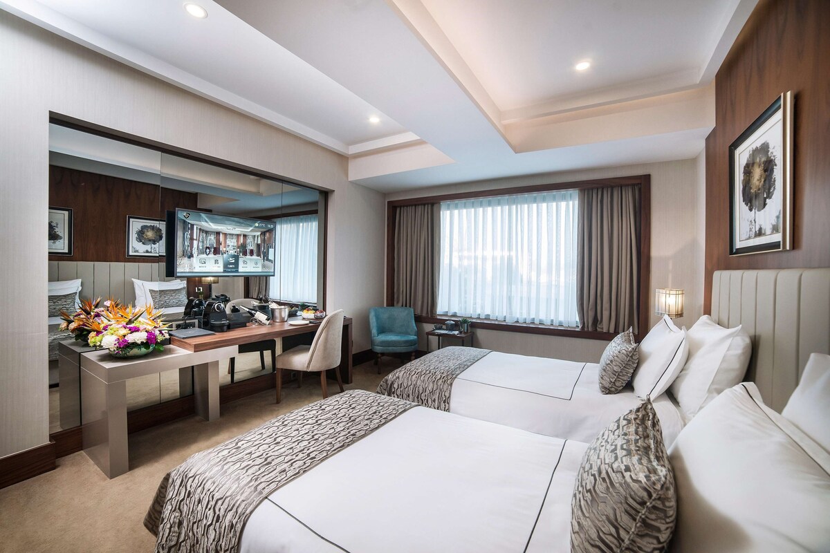 Quarto de hotel com duas camas, uma escrivaninha com computador, uma cadeira e uma janela grande com cortinas.