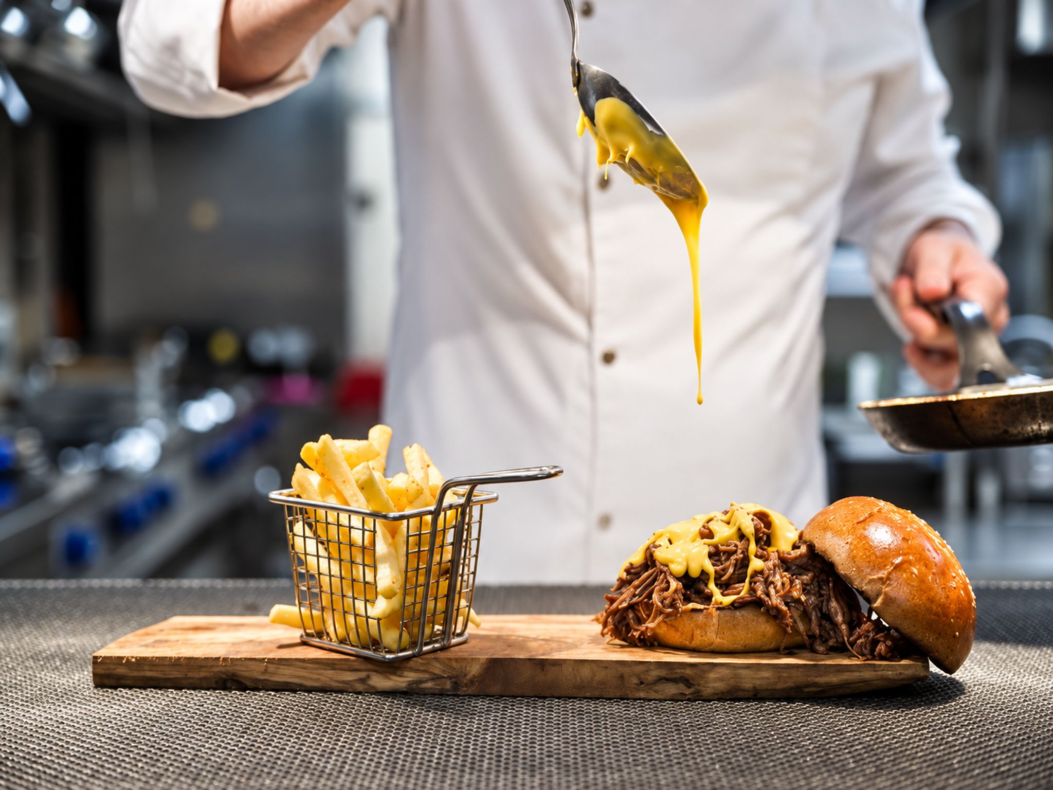 A chef holding a ladle with a yellow sauce, a basket of fries, and a pulled pork sandwich on a wooden board.