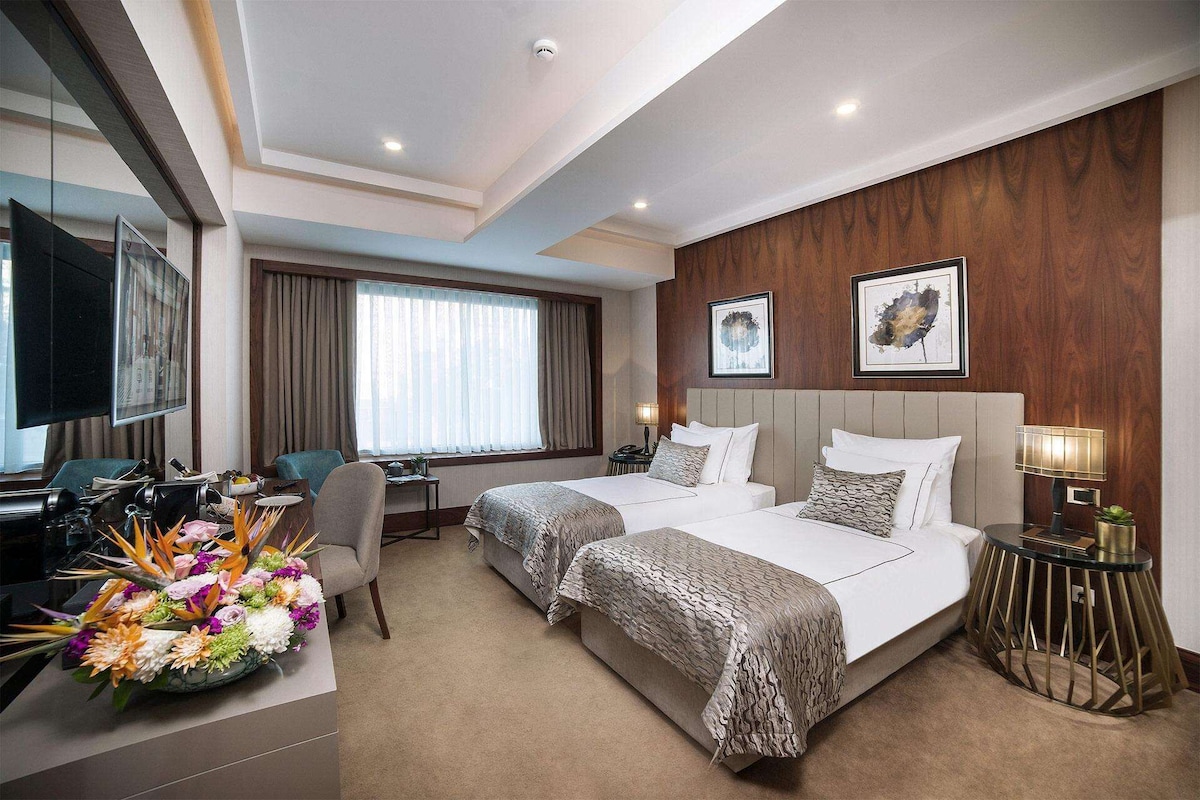 Quarto de hotel com duas camas, uma escrivaninha, uma cadeira, televisão e um arranjo floral decorativo.