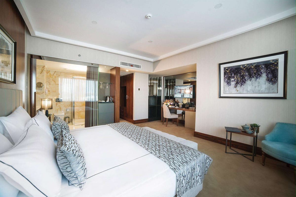 A hotel room with a large bed, a desk, a chair, a small table with a plant, and a framed artwork on the wall.