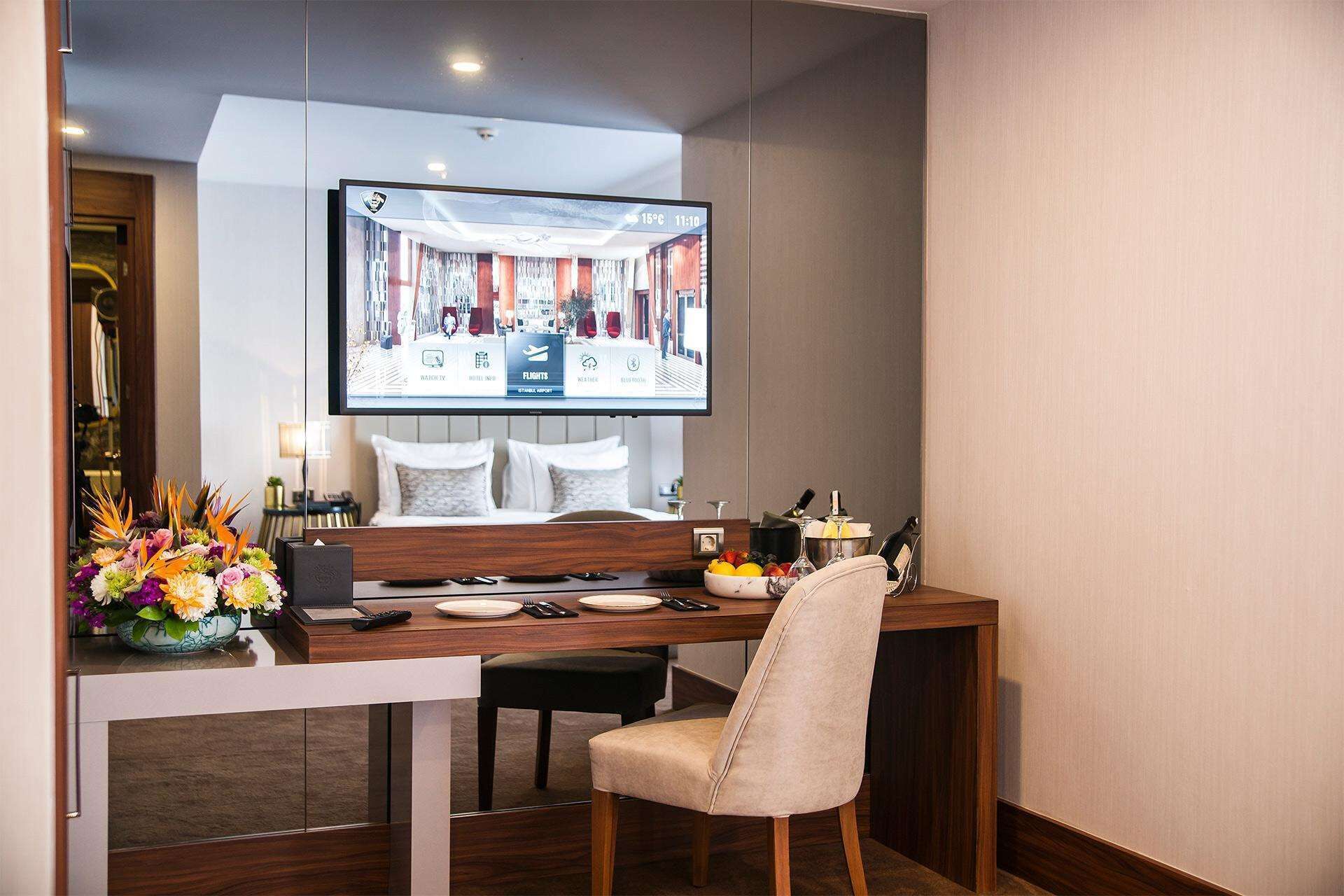 Une chambre d’hôtel comprenant un coin repas, un téléviseur diffusant des images de l’intérieur de l’hôtel, un arrangement floral et un mini-bar.