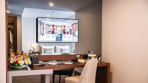 A hotel room with a dining area, a TV displaying a hotel interior, a floral centerpiece, and a minibar. - Sura Luxury Million & Spa (Istanbul)