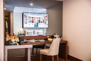 A hotel room with a dining area, a TV displaying a hotel interior, a floral centerpiece, and a minibar. - Sura Luxury Million & Spa (Istanbul)