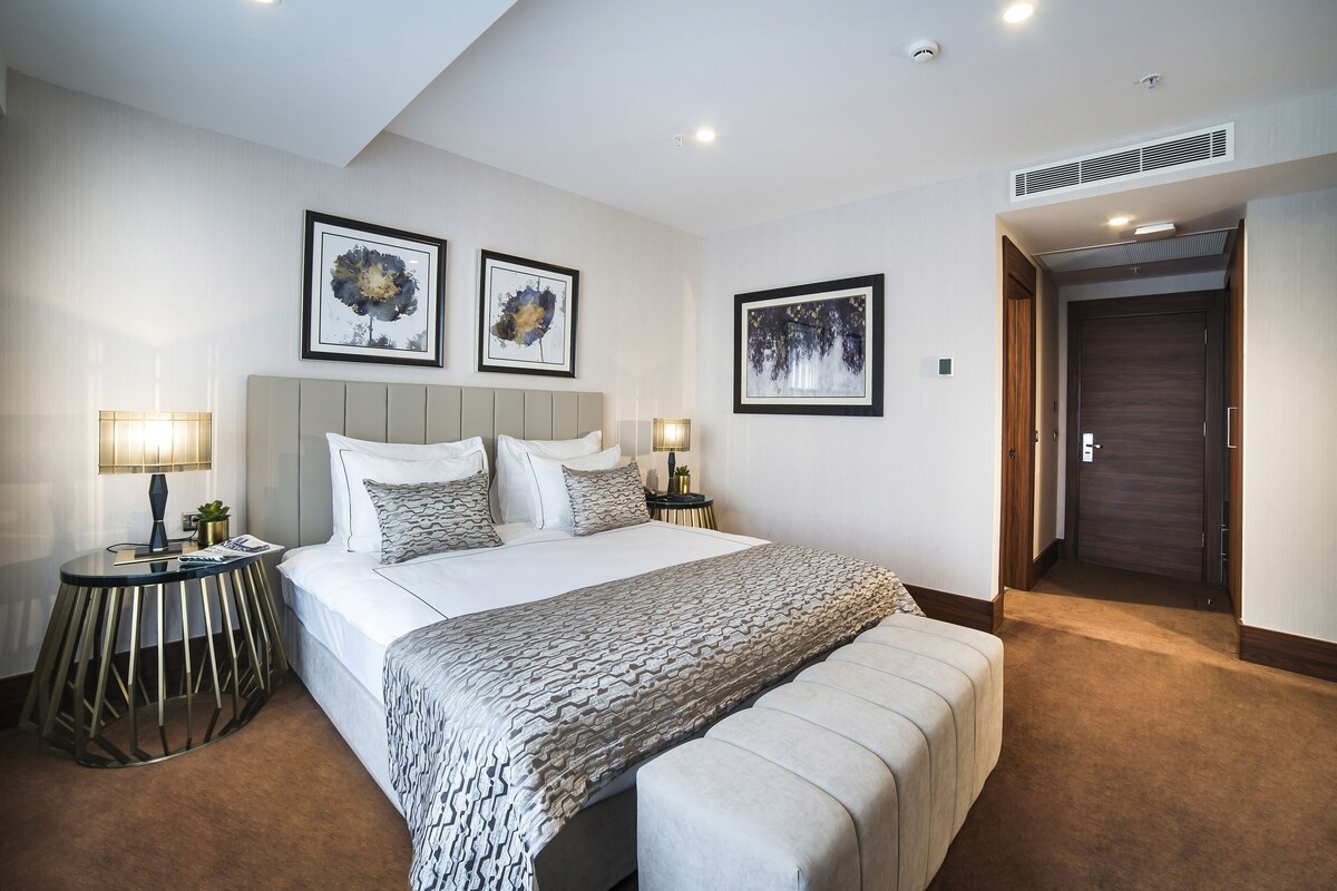 A hotel room with a large bed, bedside tables, a bench, and framed artwork on the walls.