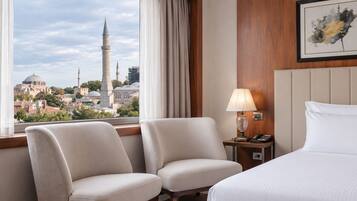 A hotel room with a bed, two armchairs, a nightstand with a lamp, and a view of a mosque and cityscape.