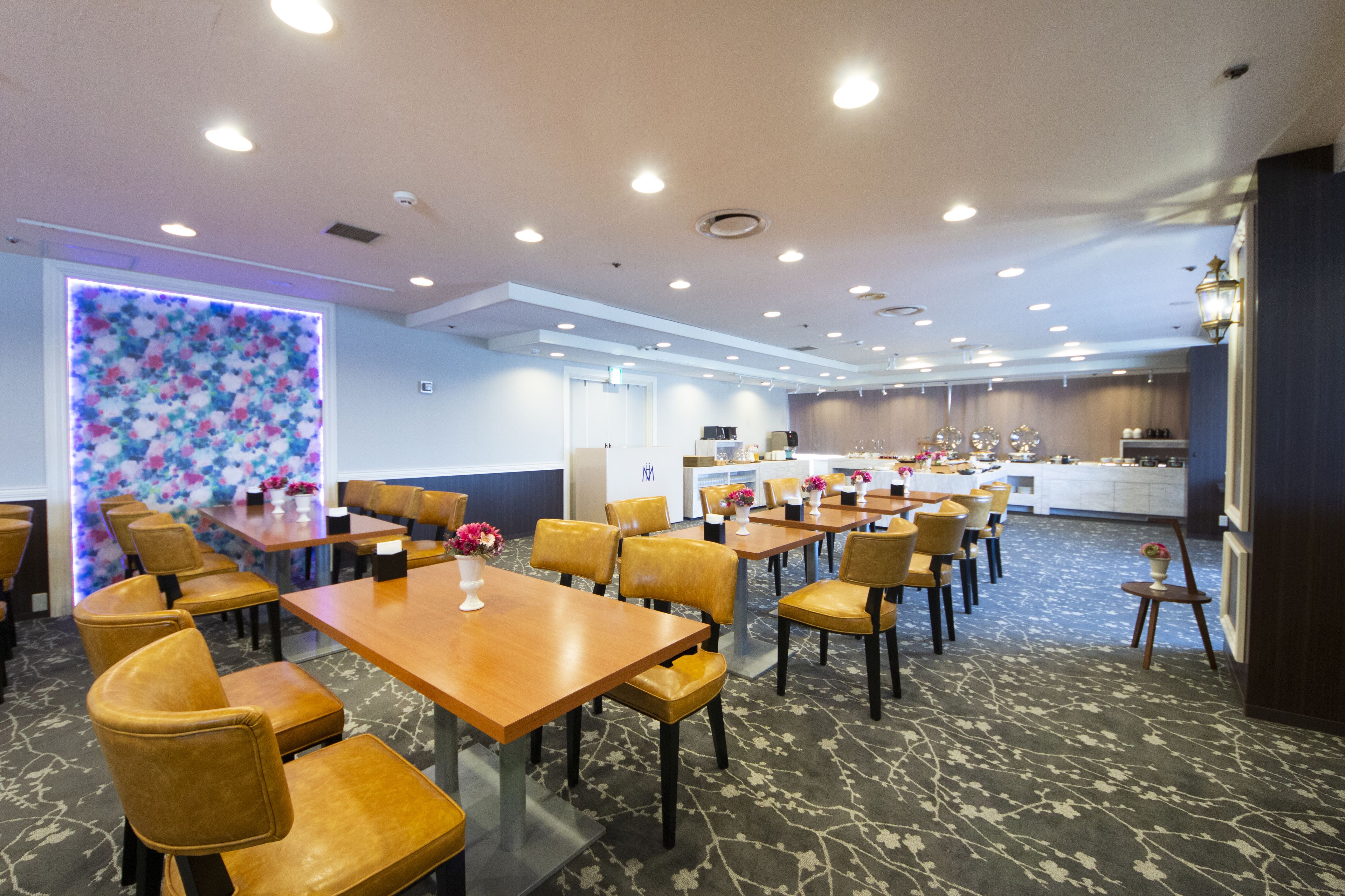 Een restaurantgedeelte in een hotel met meerdere tafels en stoelen, een keuken op de achtergrond en een decoratieve wand met een bloemistisch patroon.