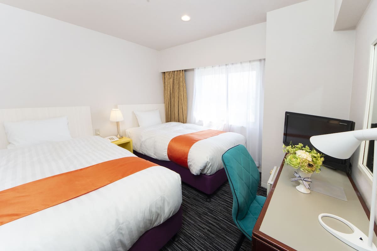 Habitación de hotel con dos camas, un escritorio con lámpara, televisor y ventana con cortinas.