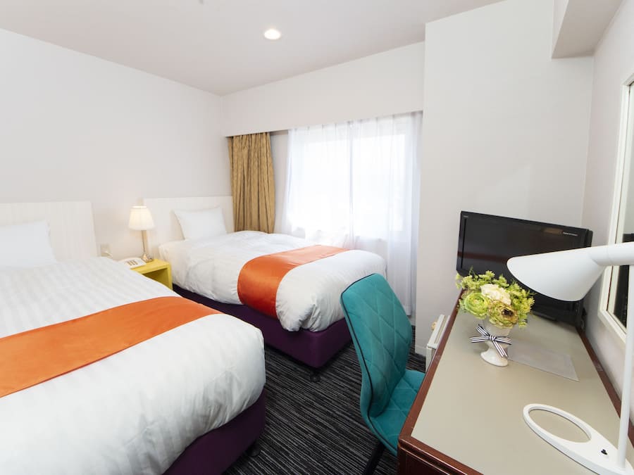A hotel room with two beds, a desk with a lamp, a TV, and a window with curtains.