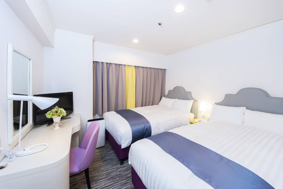 Habitación de hotel con dos camas, un escritorio con computadora y lámpara, una silla y un televisor.