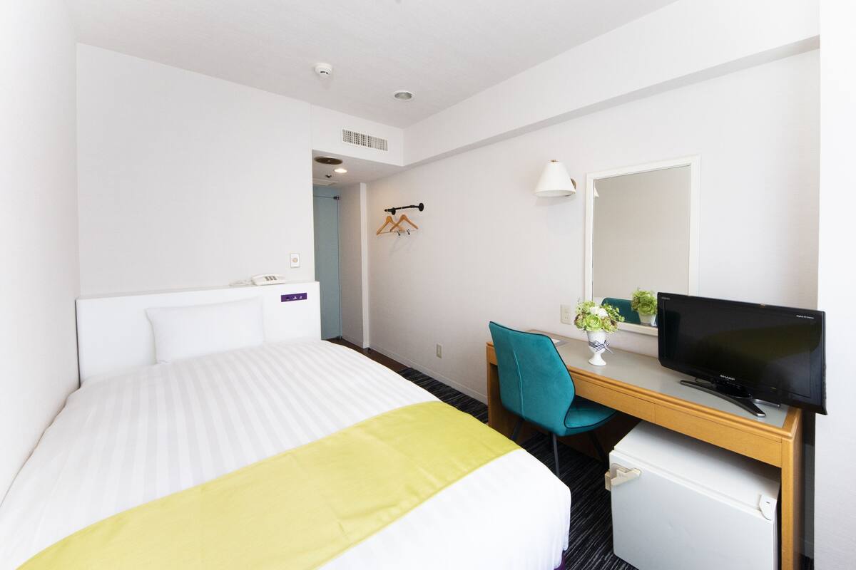 Una habitación de hotel con una cama, un escritorio con computadora, una silla y un refrigerador pequeño.