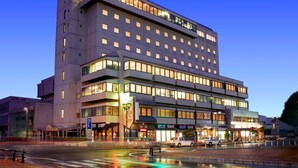 Un hotel de varios pisos con diseño moderno, que cuenta con una entrada destacada en esquina y varias tiendas a nivel de calle.