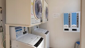 A laundry area with two washing machines and two dryers, a control panel with instructions, and a wall-mounted water softener unit. - Hotel Paco Jr Kitami (Kitami)