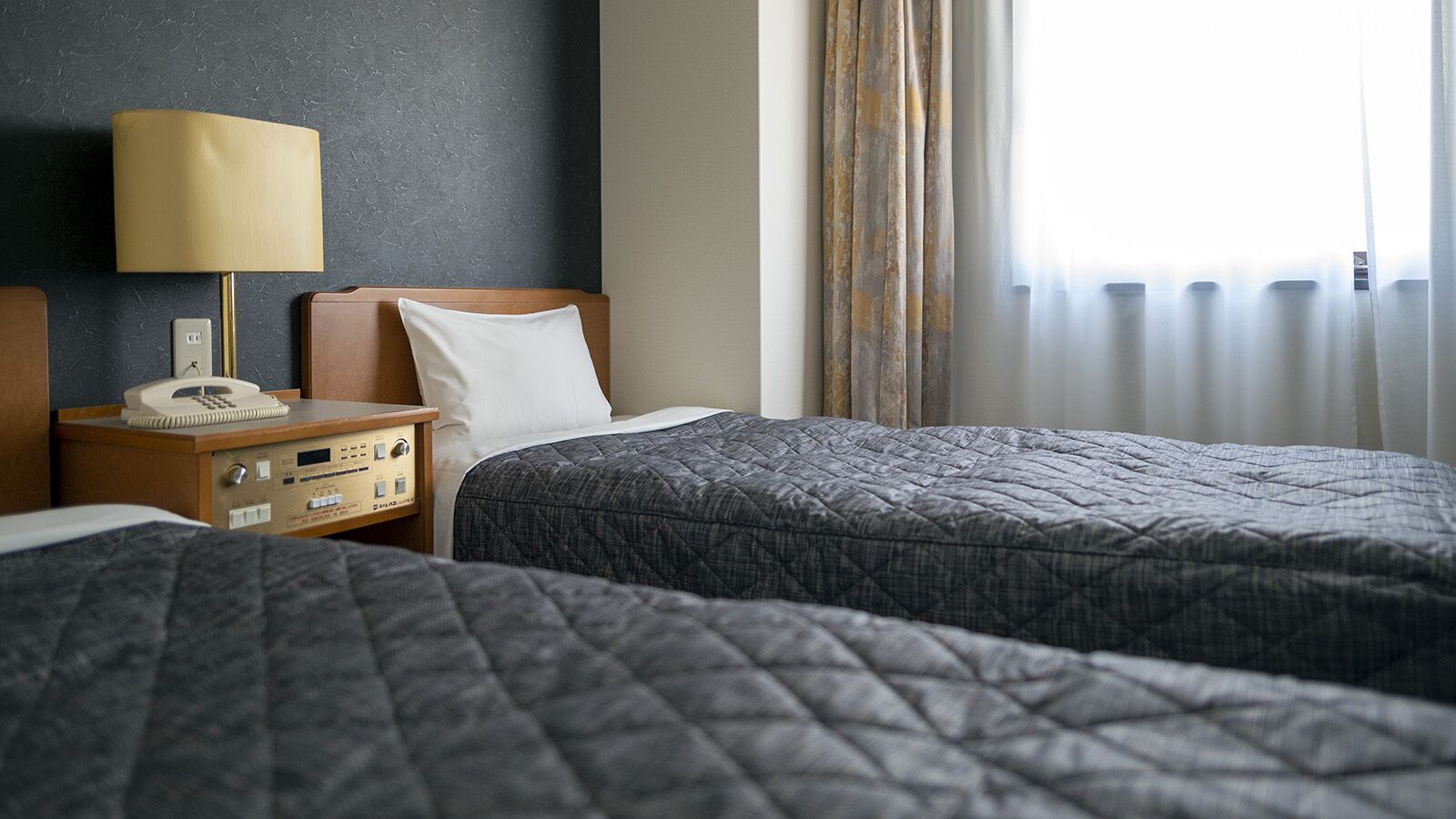 Hotellihuone, jossa on kaksi sänkyä, yöpöytä lampun kanssa, puhelin ja ikkuna, jossa on verhot.