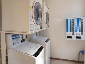 Una sala de bugaderia amb dues rentadores i una assecadora, un panell de control amb instruccions i un comptador d'aigua.